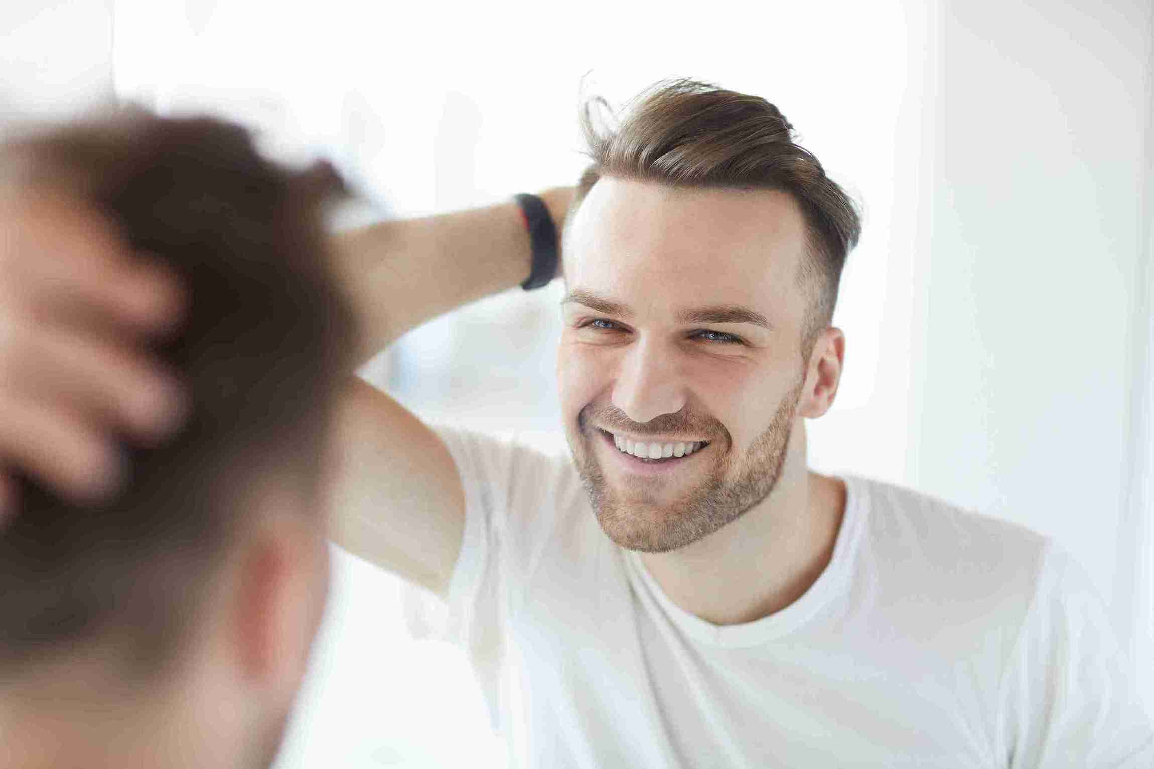 Junger Mann betrachtet lächelnd sein volles Haar im Spiegel – Symbol für eine hohe Haartransplantation Erfolgsquote.