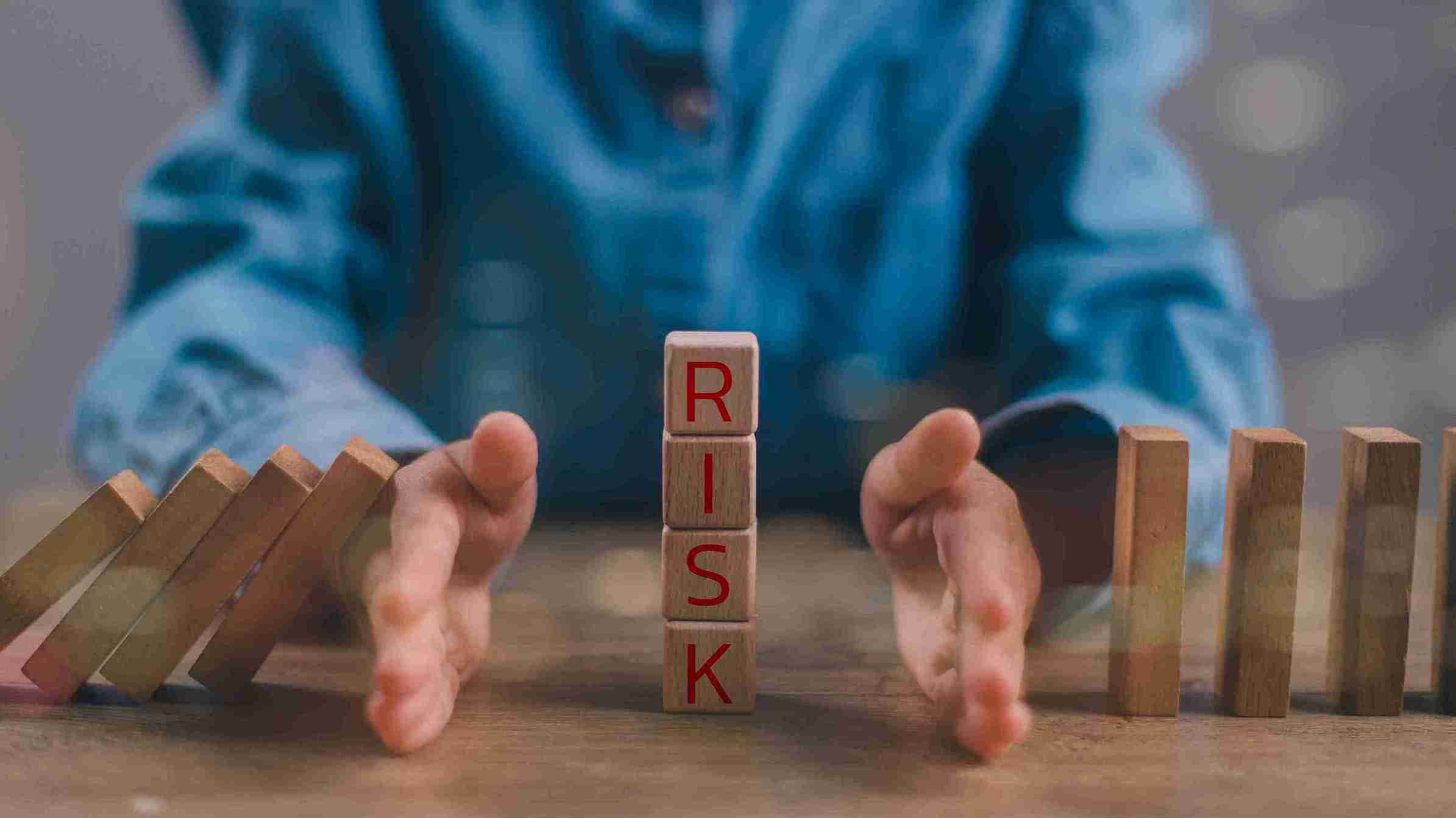 A person in a blue shirt uses their hands to stop falling dominoes, with wooden blocks stacked vertically spelling the word 'RISK' in red letters, symbolizing prevention and awareness of hair transplant risks.
