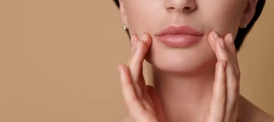 Gros plan sur le bas du visage d'une femme aux lèvres pulpeuses et harmonieuses, encadrant délicatement son menton avec ses mains après un traitement par injection de produit de comblement des lèvres.
